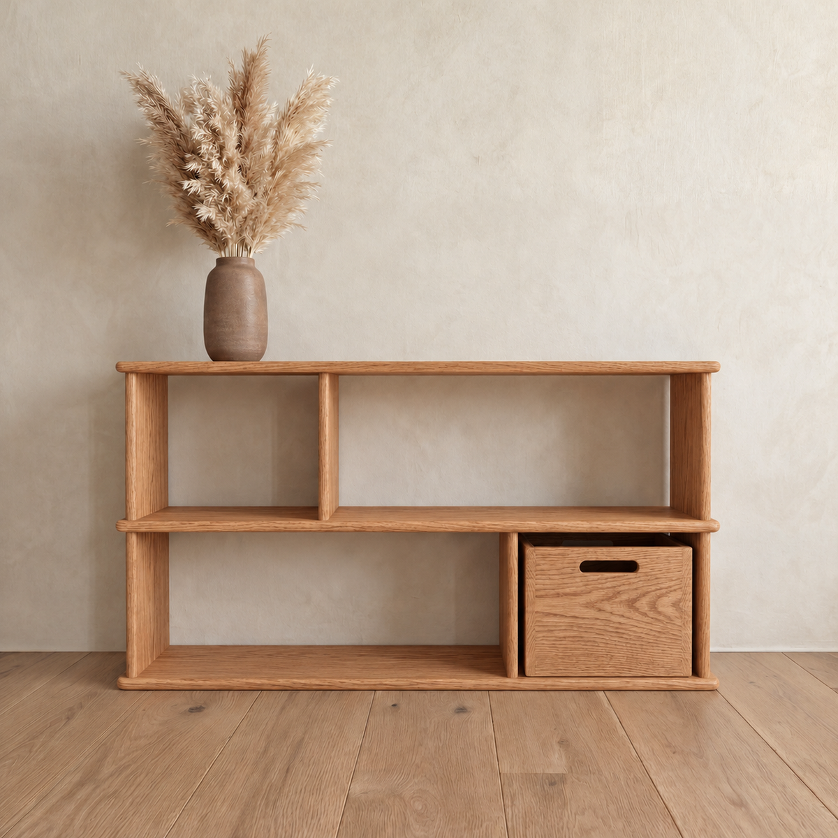 The horizontal bookshelf in red oak against a plaster wall, with a vase of pampas grass on top and a wooden bin in one of the lower openings.