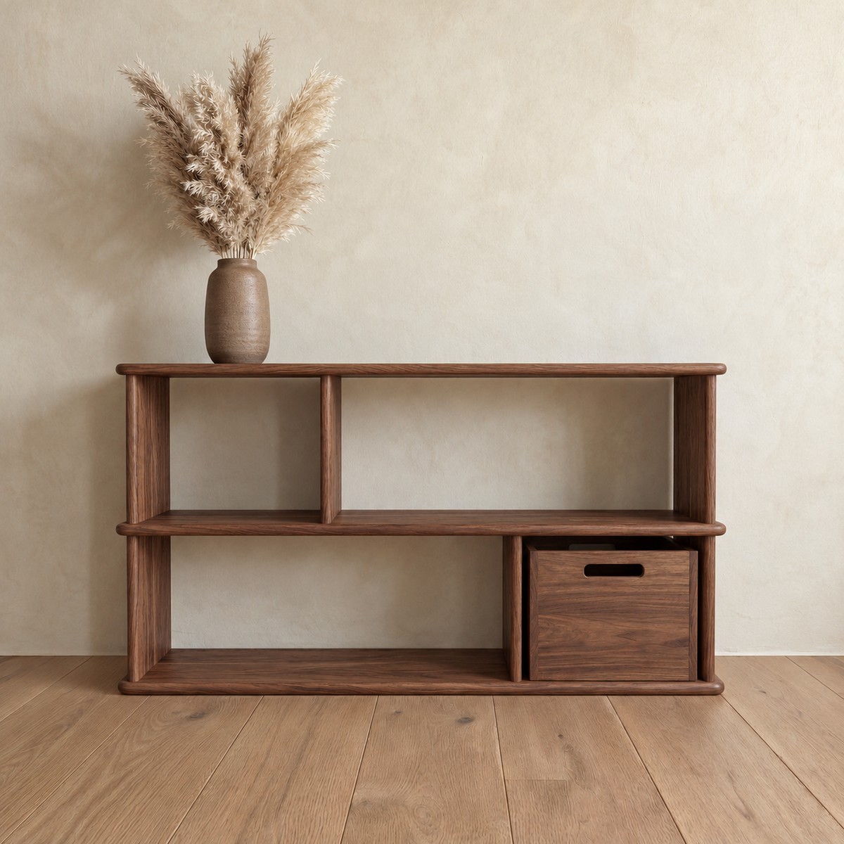 The horizontal bookshelf in walnut against a plaster wall, with a vase of pampas grass on top and a wooden bin in one of the lower openings.
