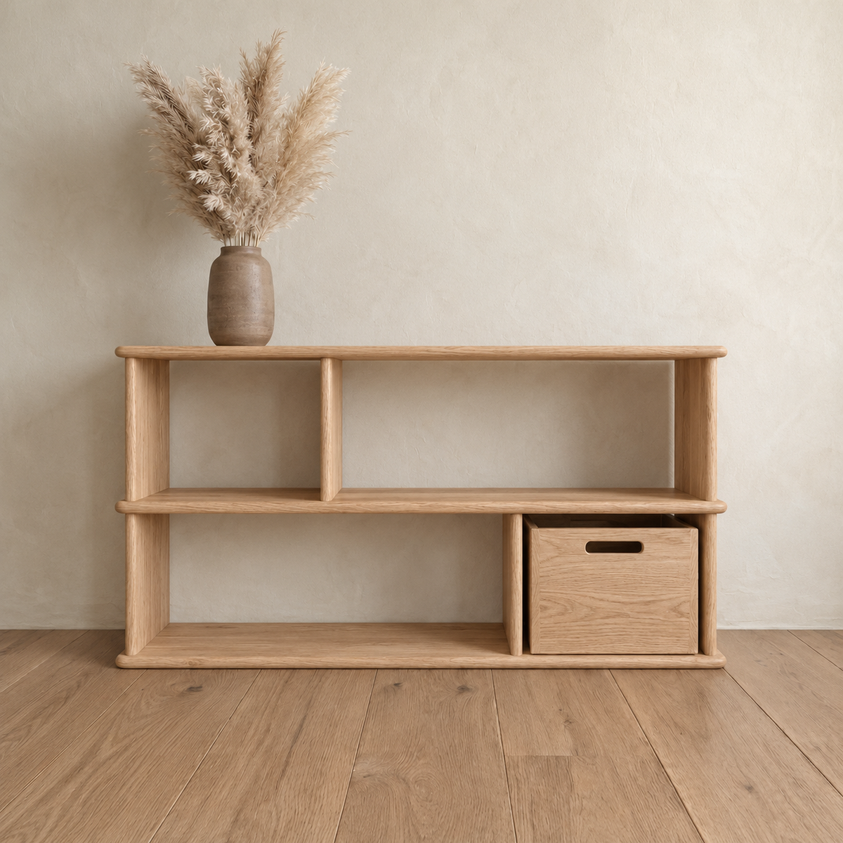 The horizontal bookshelf in white oak against a plaster wall, with a vase of pampas grass on top and a wooden bin in one of the lower openings.