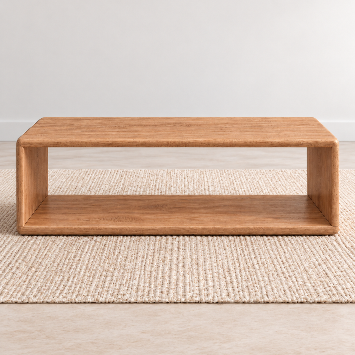 A Japandi coffee table in red oak: a low, open rectangle with softly rounded corners and a lower shelf, on a woven jute rug against a plain light wall.