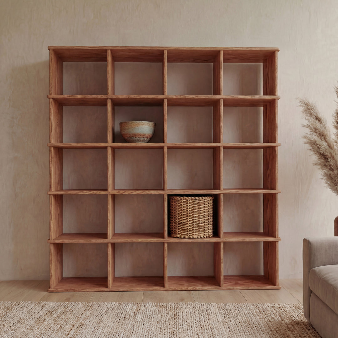 A Cube bookshelf in red oak with a 5x4 grid stands against a beige wall, holding a ceramic bowl and woven basket on mostly empty shelves. A beige rug and pampas grass are nearby.