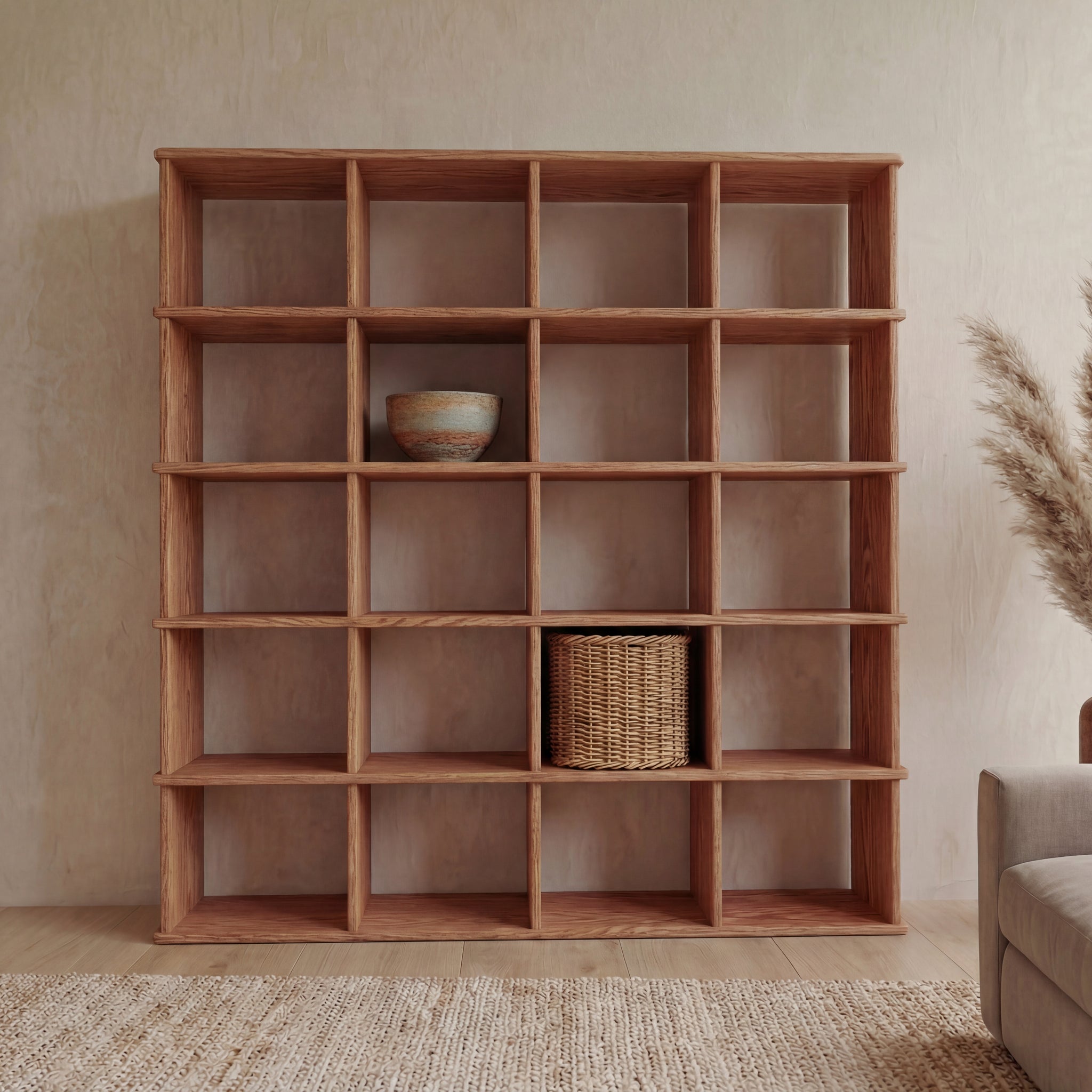 A Cube bookshelf in red oak with a 5x4 grid stands against a beige wall, holding a ceramic bowl and woven basket on mostly empty shelves. A beige rug and pampas grass are nearby.