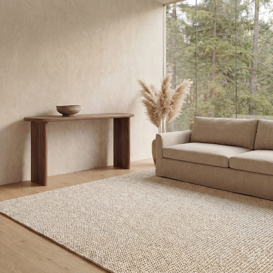 A minimalist living room features a beige sofa, textured rug, and a 60 inch console table in walnut handmade in Vancouver, accented with a bowl and tall vase of pampas grass. Floor-to-ceiling windows showcase tree views outside.