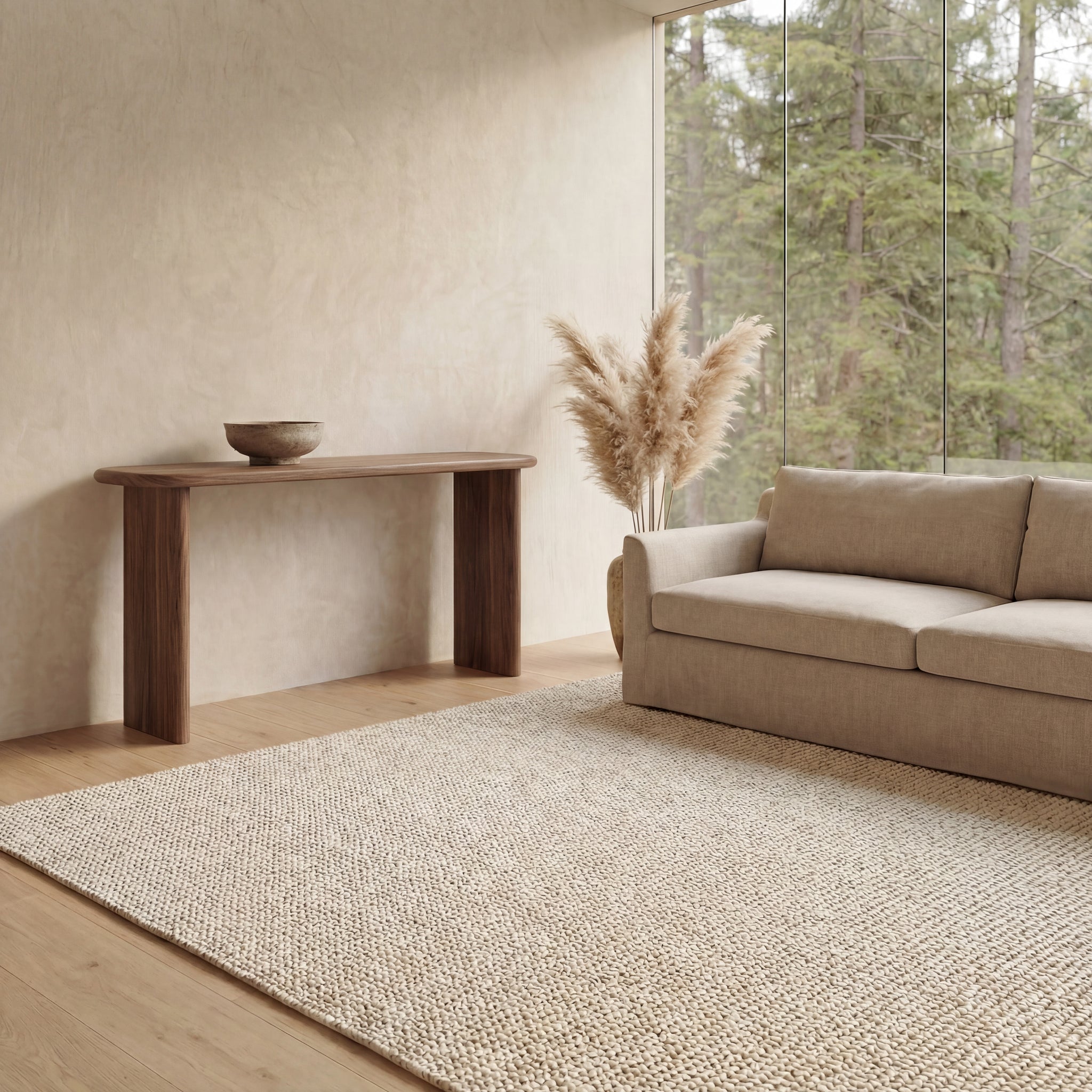 A minimalist living room features a beige sofa, textured rug, and a 60 inch console table in walnut handmade in Vancouver, accented with a bowl and tall vase of pampas grass. Floor-to-ceiling windows showcase tree views outside.