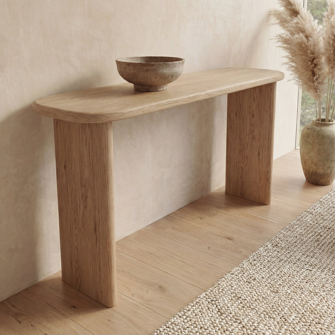 A 60 inch console table in white oak with a round bowl on top stands against a beige wall. To the right, a large vase filled with pampas grass sits on the wooden floor near a textured rug.