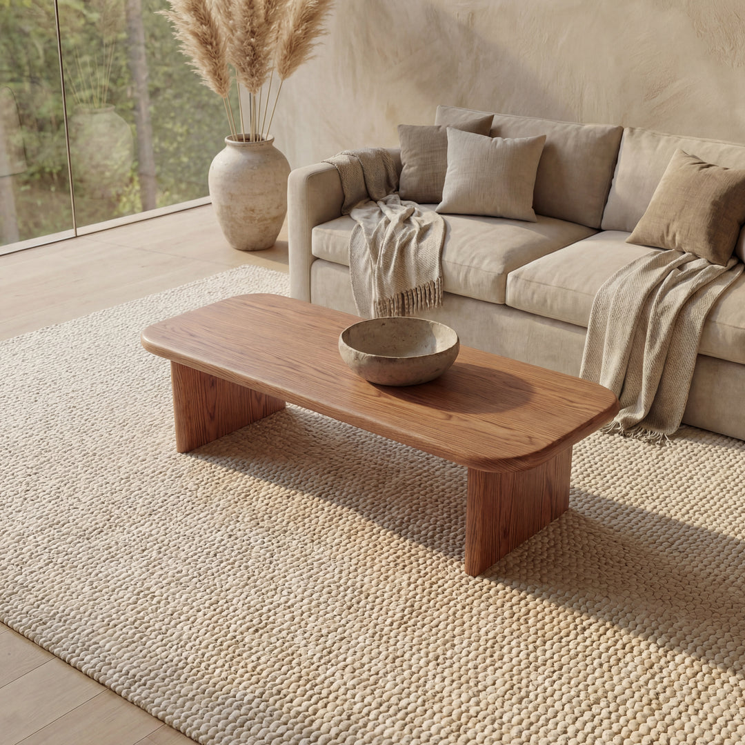 A minimal living room with a beige sofa, neutral cushions, a throw blanket, and a 60 inch coffee table in red oak topped with a ceramic bowl on a textured rug. A large vase with pampas grass stands near floor-to-ceiling windows.