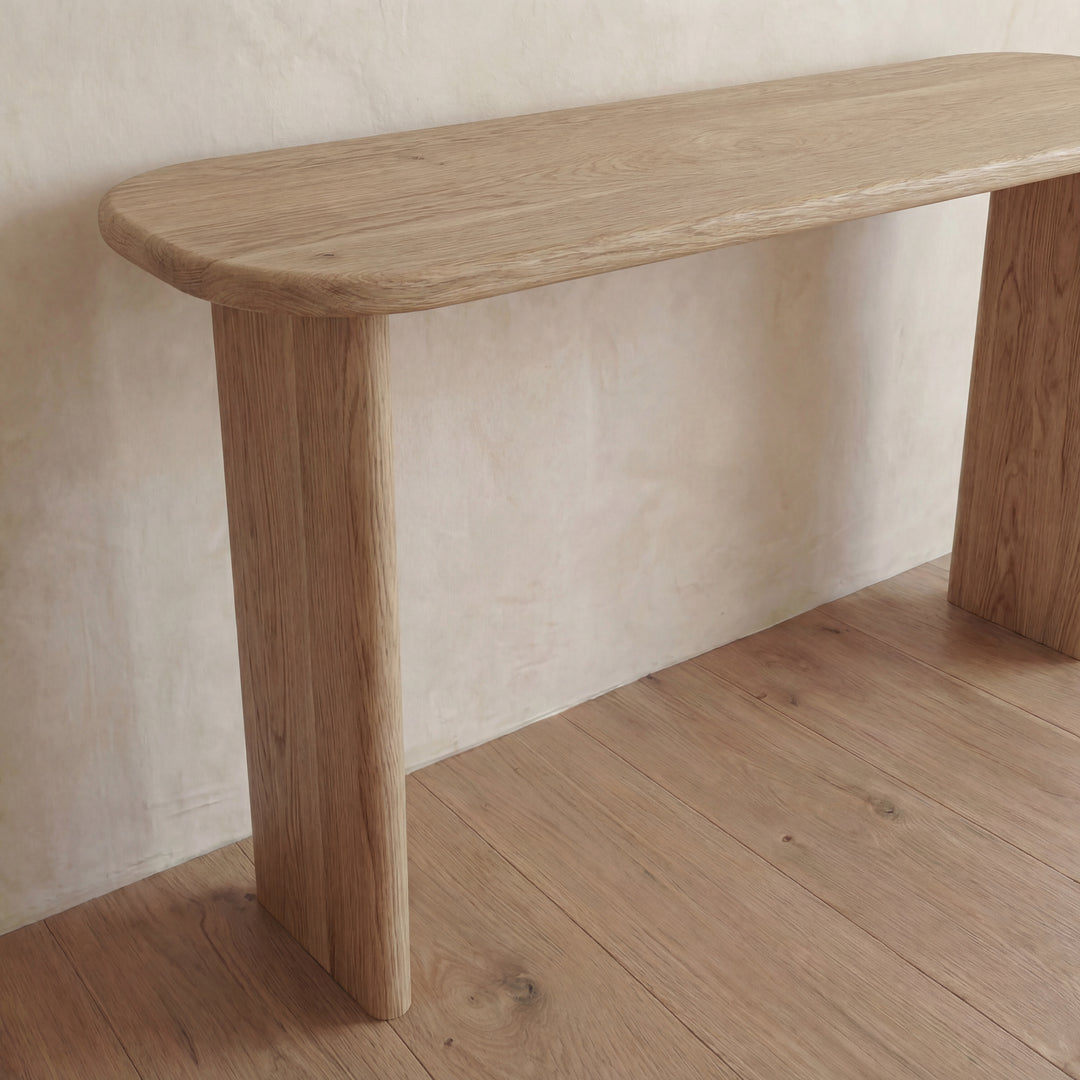The 60 inch console table in white oak, with rounded edges and sturdy legs, sits gracefully against a light-colored wall atop a matching wooden floor.