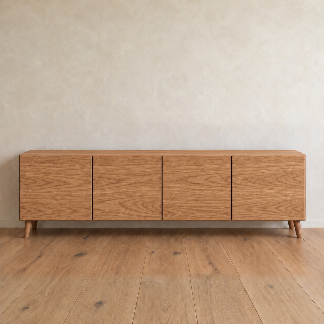 A media console in red oak with four flat doors and tapered legs, against a plain plaster wall on a wood floor.