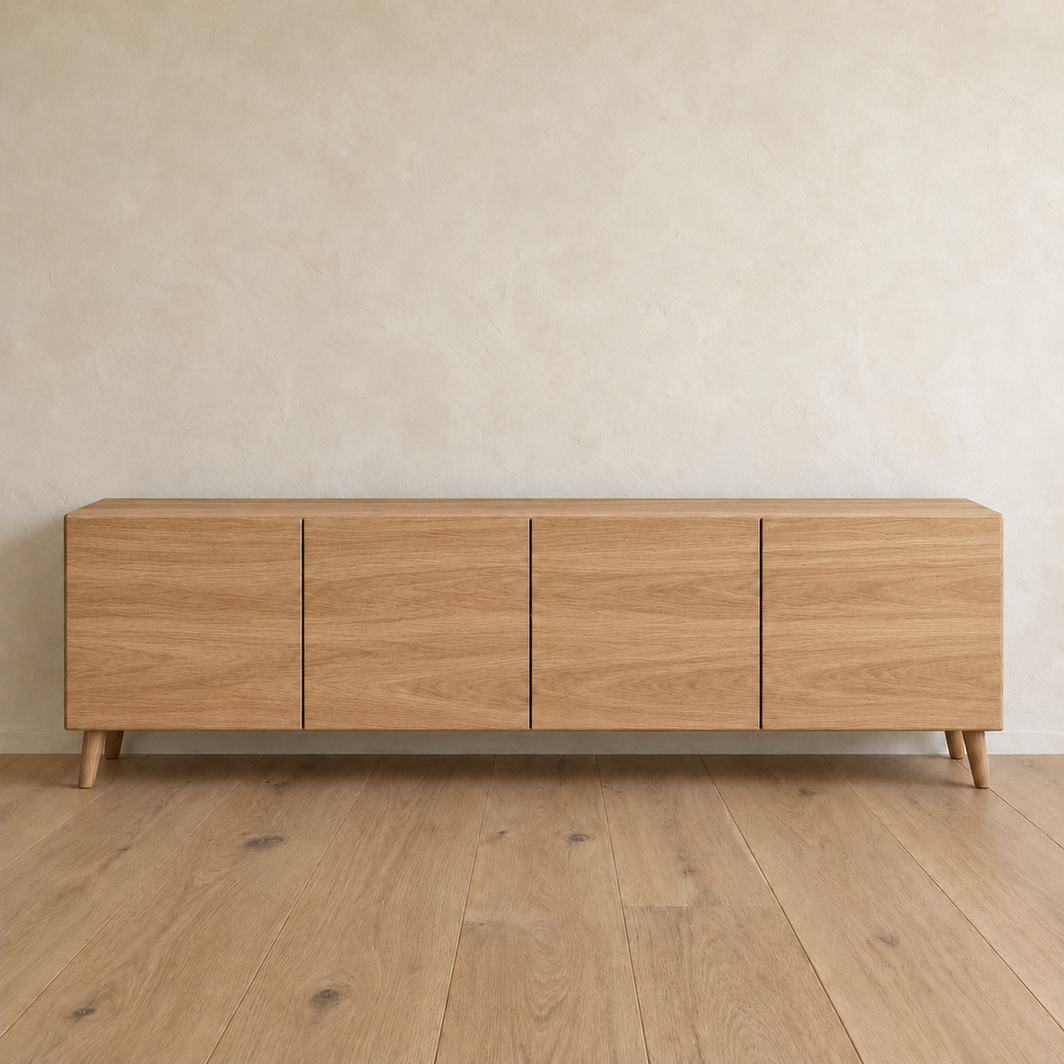 A media console in white oak with four flat doors and tapered legs, against a plain plaster wall on a wood floor.