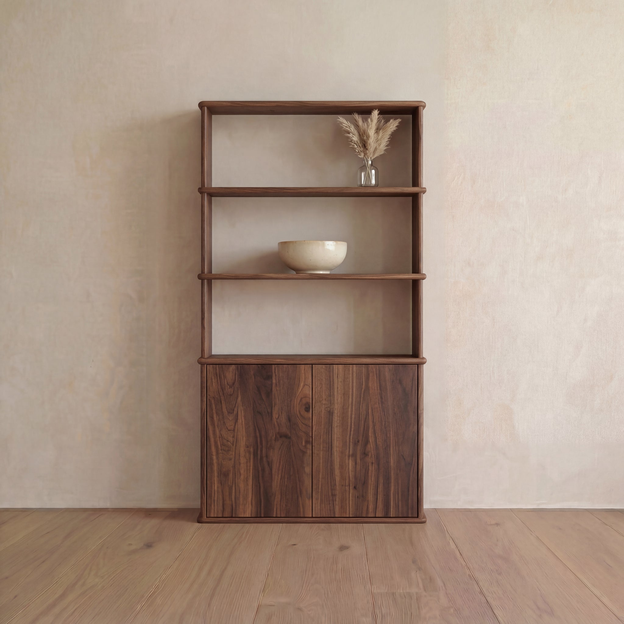 The Cabinet bookshelf in walnut features a minimalist design with three open shelves and a push-to-open cabinet at the base. A ceramic bowl is displayed on the middle shelf, while a glass vase filled with dried grass decorates the top shelf, all set against a neutral wall backdrop.