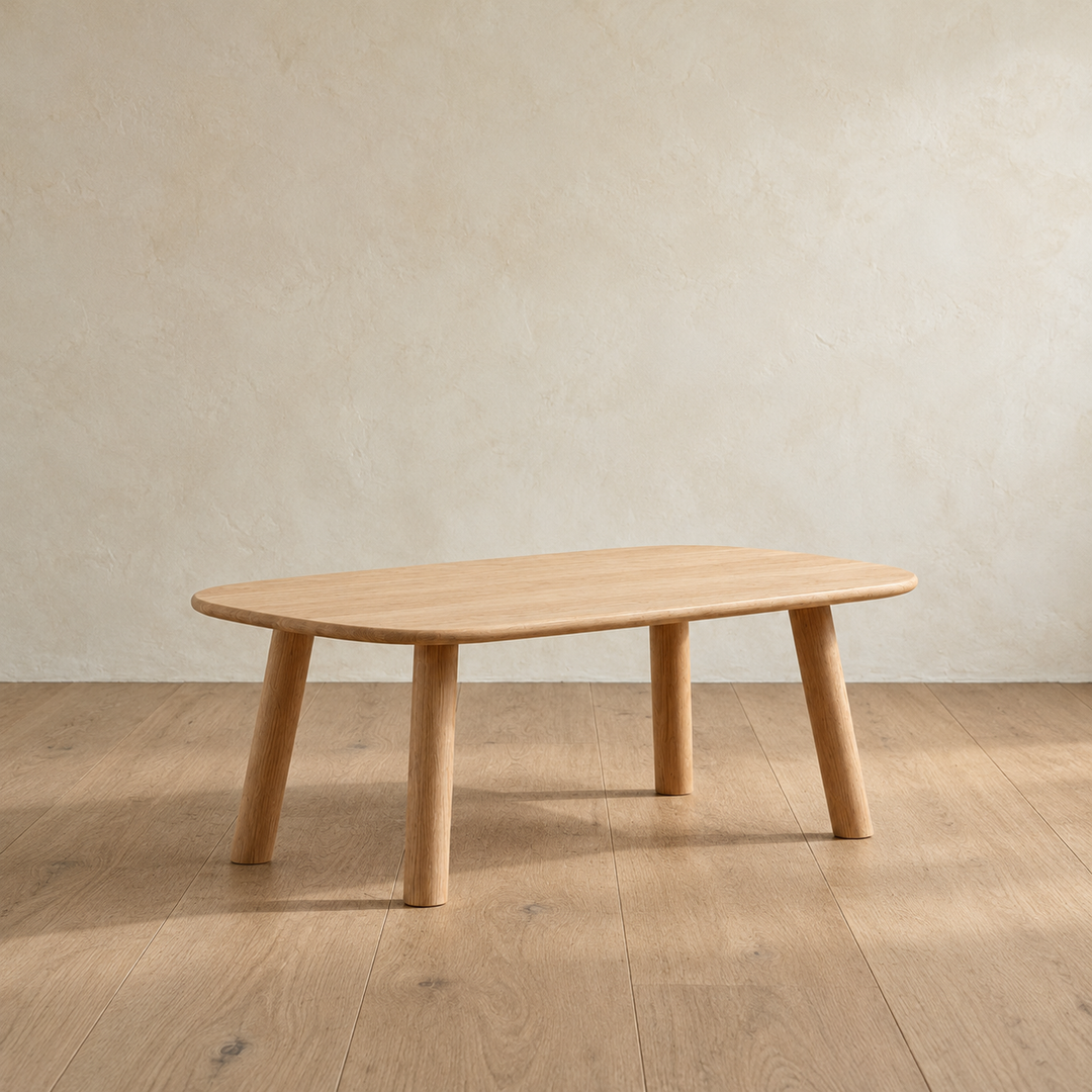 A minimalist coffee table in white oak with a slim top, softly rounded corners and four splayed round legs, against a plain plaster wall.
