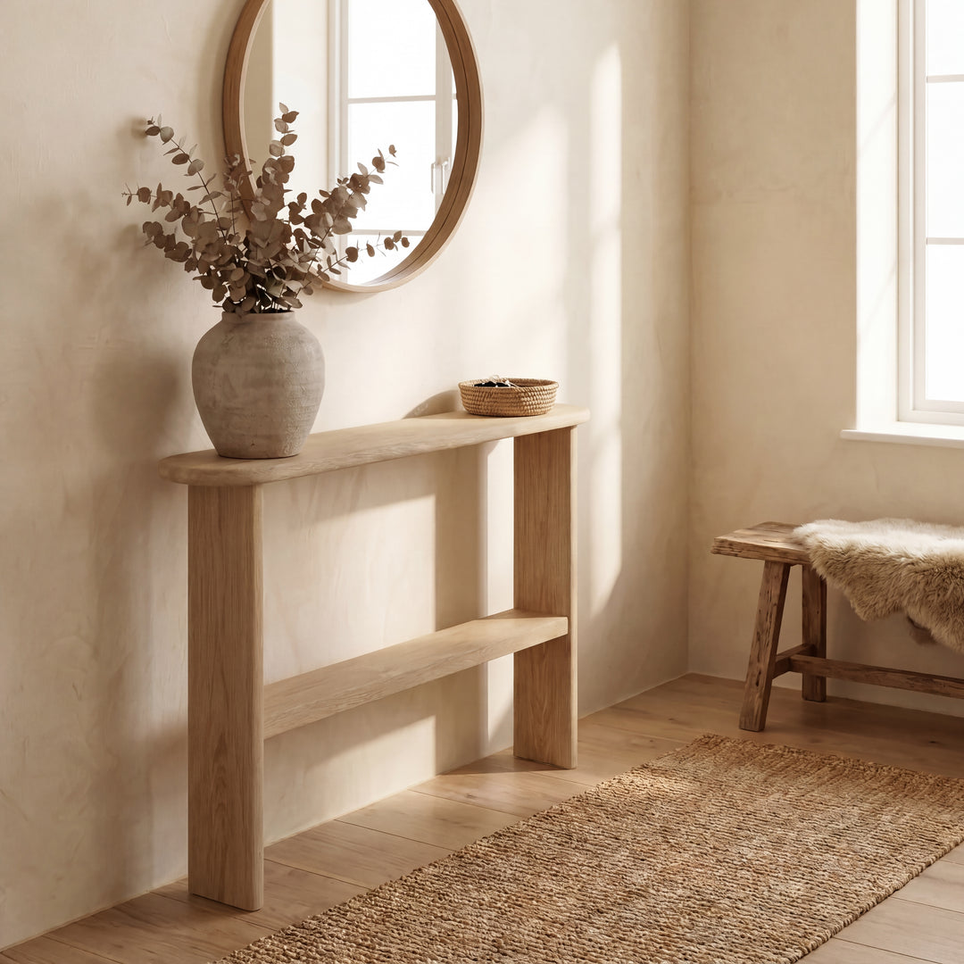 A minimalist entryway features a handmade console table with shelf in white oak, a round mirror above it, a large ceramic vase with dried branches, a small woven basket, a wooden bench draped with a fur throw, and sunlight streaming through the window.