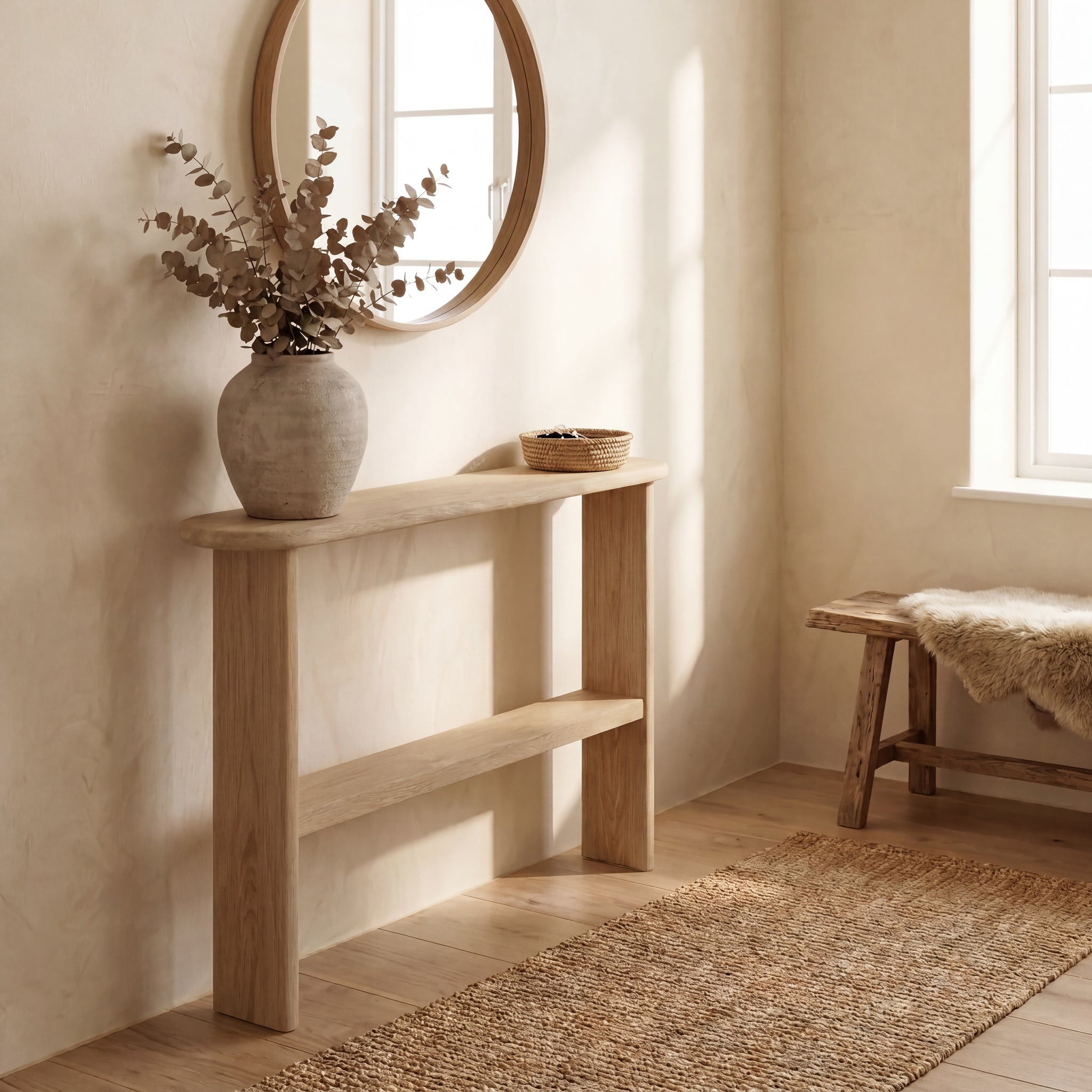 A minimalist entryway features a handmade console table with shelf in white oak, a round mirror above it, a large ceramic vase with dried branches, a small woven basket, a wooden bench draped with a fur throw, and sunlight streaming through the window.