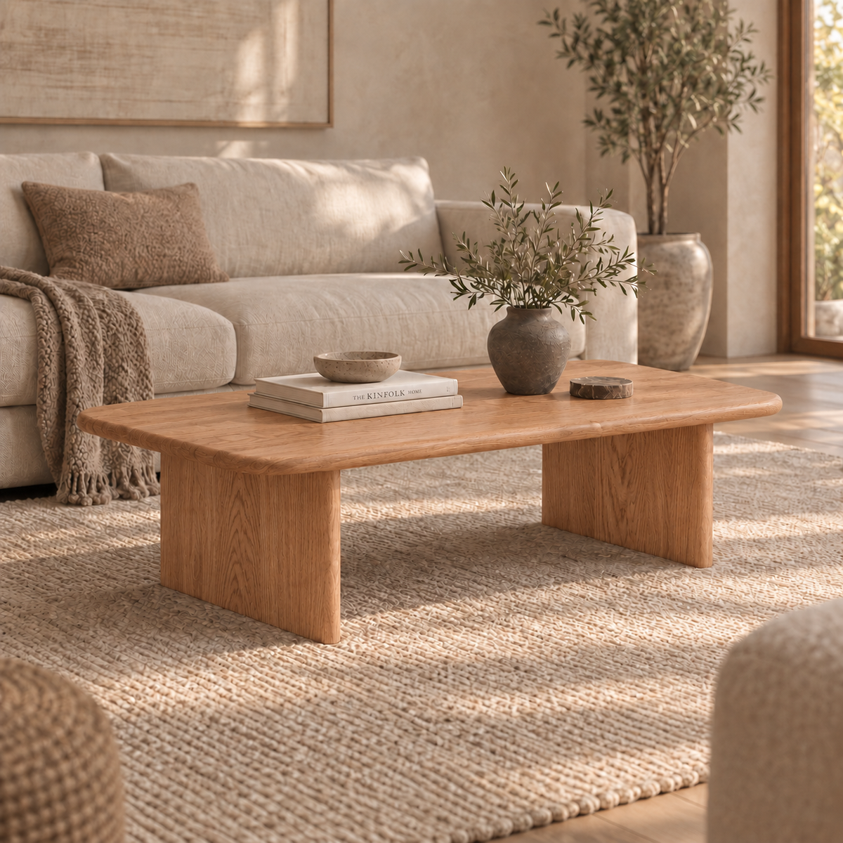 Minimalist living room with a light beige sofa, textured pillows, and a 60 inch coffee table in red oak holding books and a vase, complemented by a large woven rug, neutral décor, and soft natural light from windows.