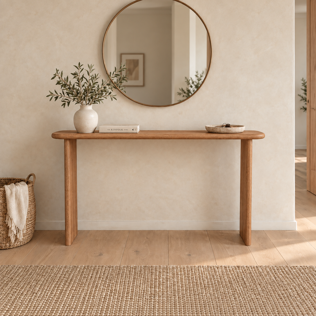 The Narrow Console Table in Red Oak, handmade in Vancouver, stands against a beige wall beneath a round mirror. On its surface rest a vase of greenery, a book, and a bowl. Nearby are a woven basket holding a blanket and a textured rug.