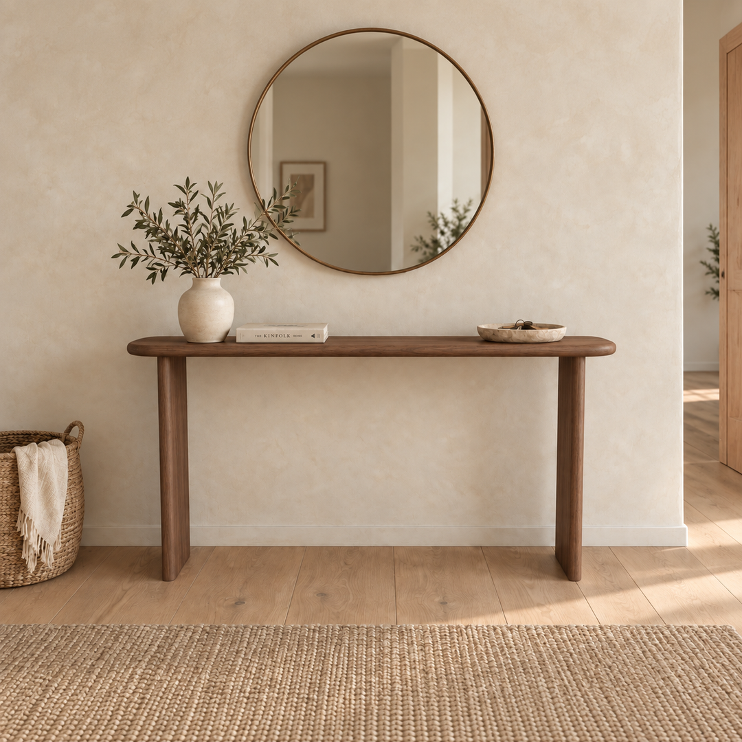 A minimalist entryway featuring a narrow console table in walnut, a round wall mirror, a vase with green branches, a shallow bowl, a book, a woven basket, and neutral decor on light wood floors.