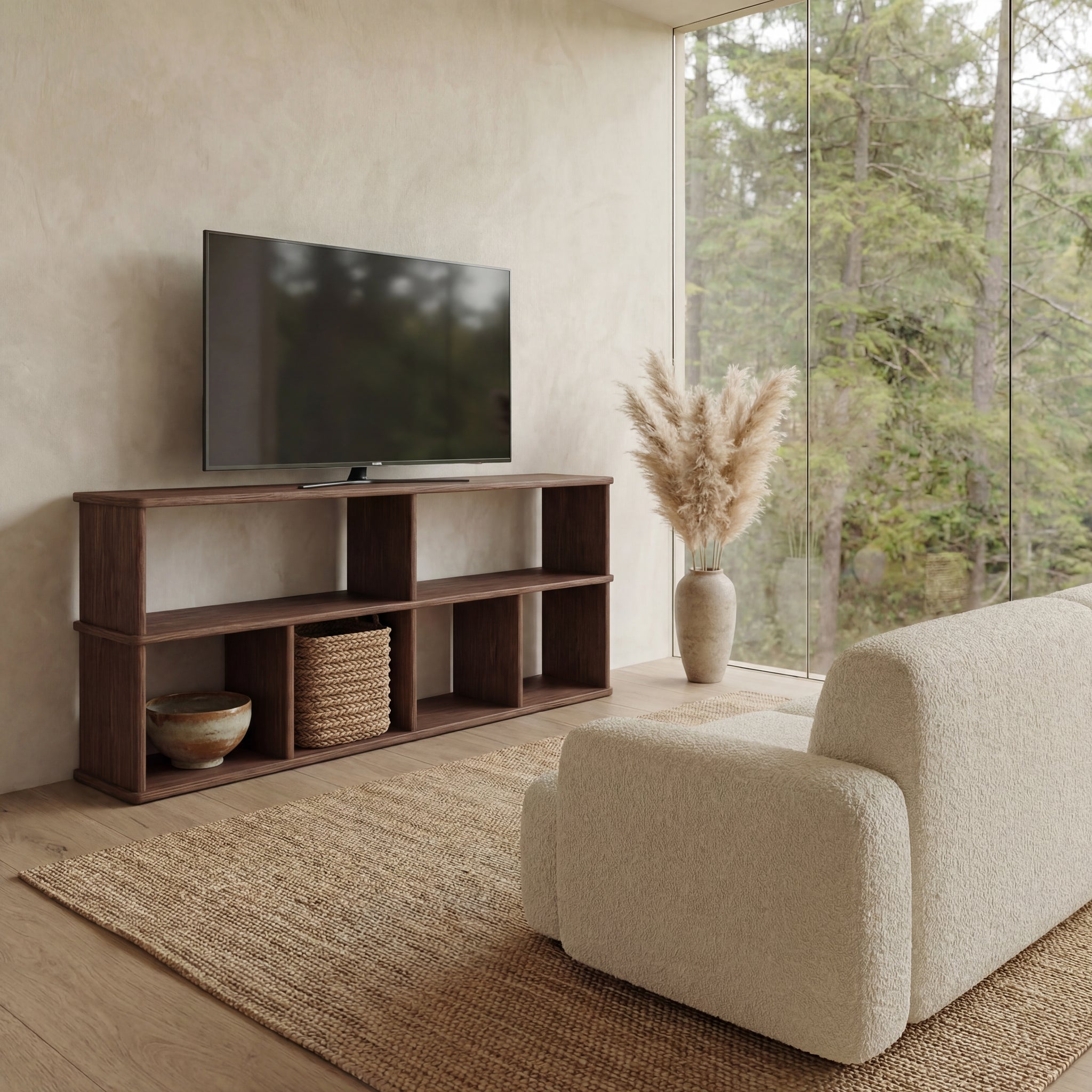 A minimalist living room with a beige sofa, 65 inch bookshelf TV stand in walnut, flat-screen TV, large vase with pampas grass, woven basket, and floor-to-ceiling windows overlooking trees.