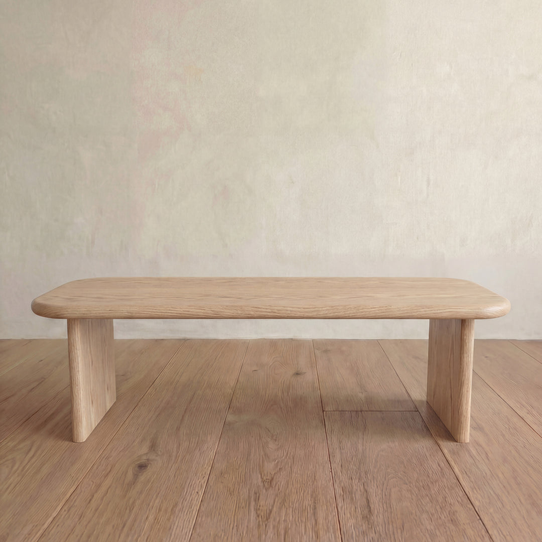 A 60 inch coffee table in white oak with a minimalist design and rounded edges sits on a wooden floor against a plain, light-colored wall.