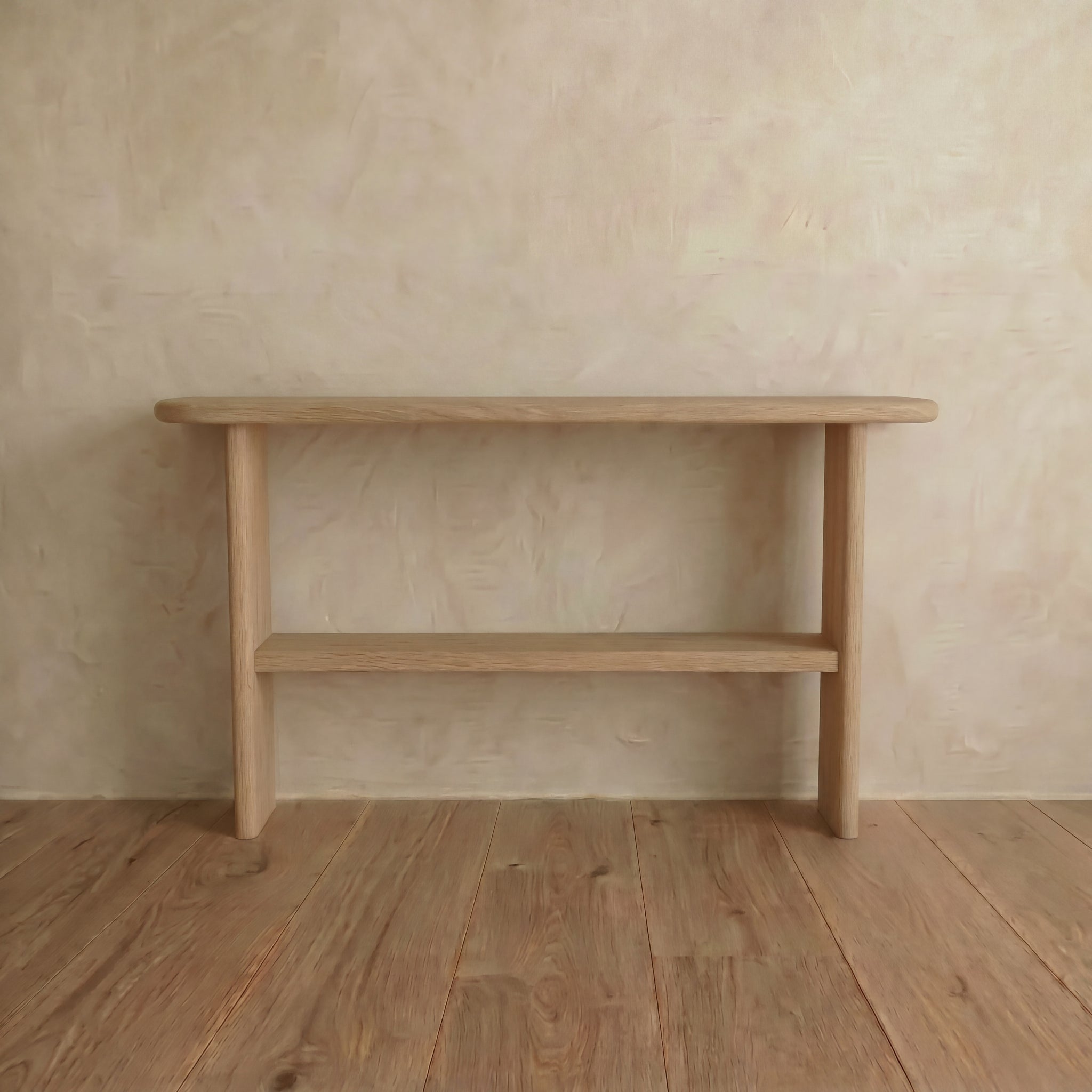 The Console table with shelf in white oak features a minimalist handmade design, rounded edges, and light wood grain. It stands against a textured beige wall on a wooden floor, with a convenient lower shelf for extra storage.