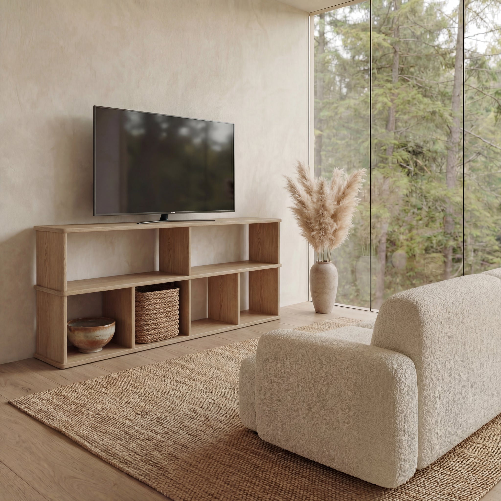 A modern living room with a beige sofa, a 65 inch bookshelf TV stand in white oak holding a flatscreen TV, a woven rug, and pampas grass in a vase by floor-to-ceiling windows overlooking trees.