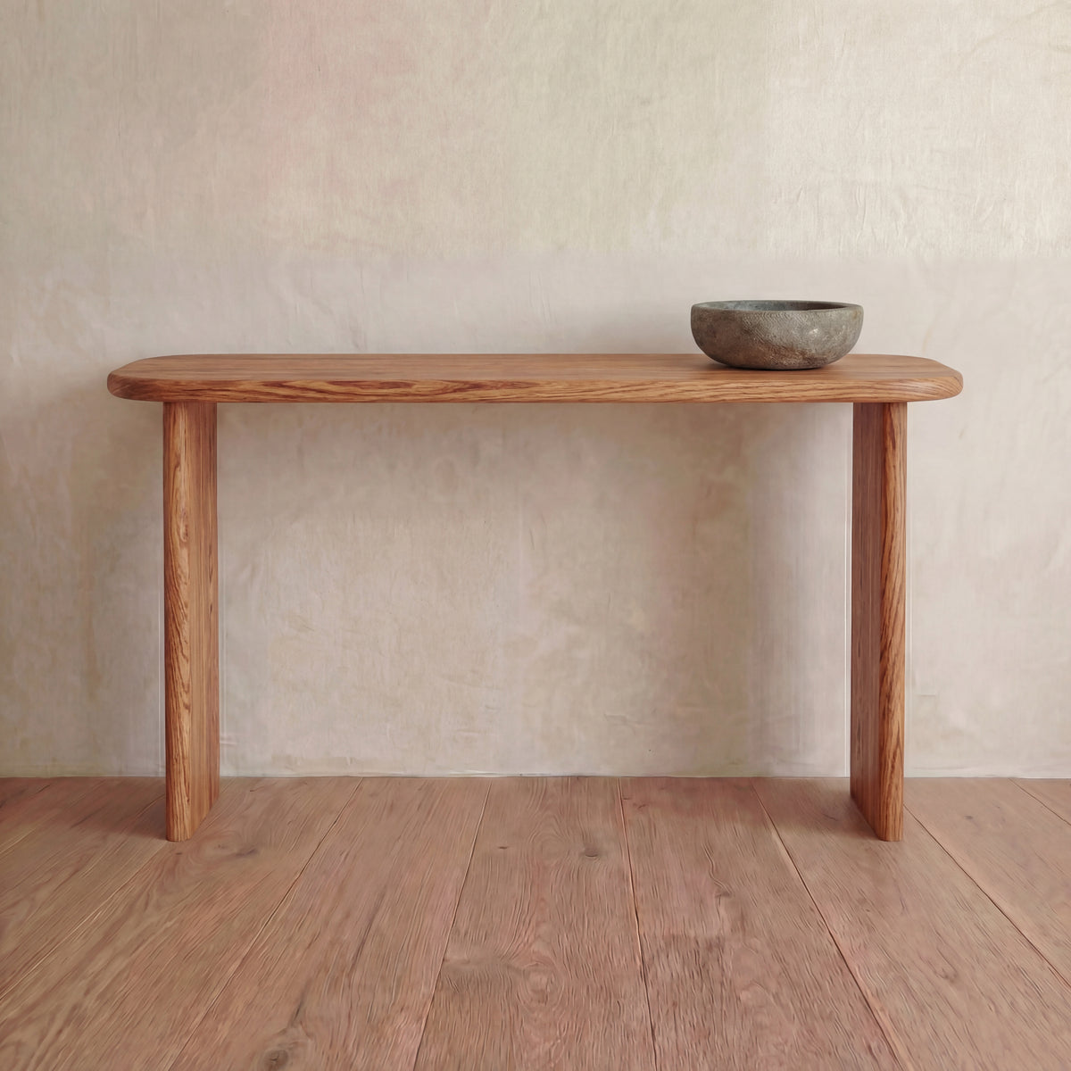 A 60 inch red oak console table with rounded edges sits against a light textured wall, topped with a single gray stone bowl. The table and floor feature matching wood grain.