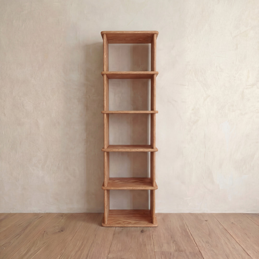 The Narrow bookshelf in red oak features five empty shelves and a tall, slim handmade design. It stands against a light beige textured wall on a wooden floor.