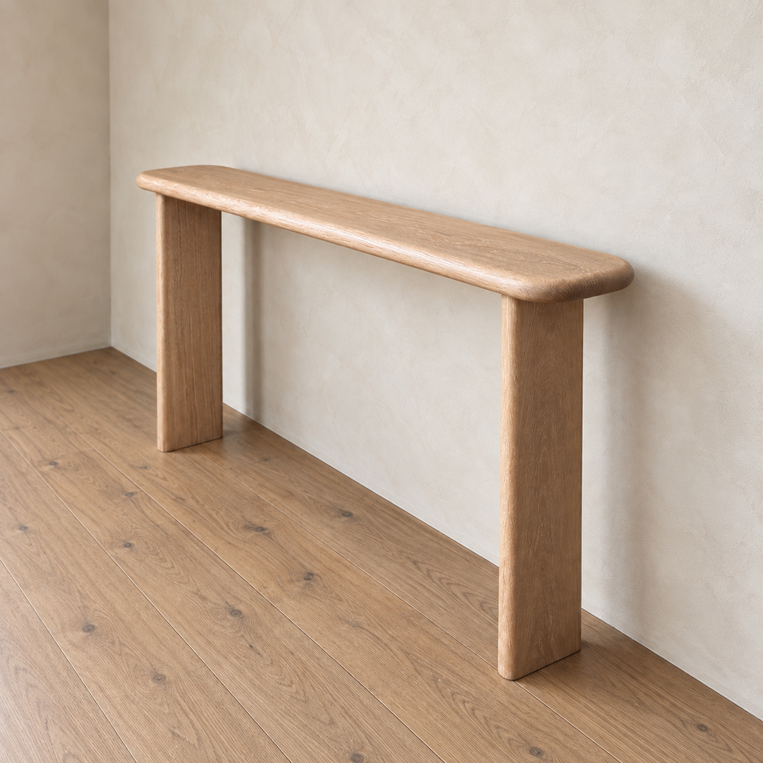 A simple and light Narrow console table in white oak with rounded edges and two thick legs stands against a beige textured wall on a wooden floor in a minimalist room. Expertly handmade in Vancouver, this narrow console table brings understated elegance to any space.