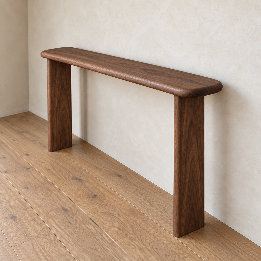 A narrow console table in walnut, crafted with a minimalist and modern design, stands against a light textured wall on a wooden floor. Handmade from solid walnut, it features rounded edges, two thick legs, and a smooth finish.