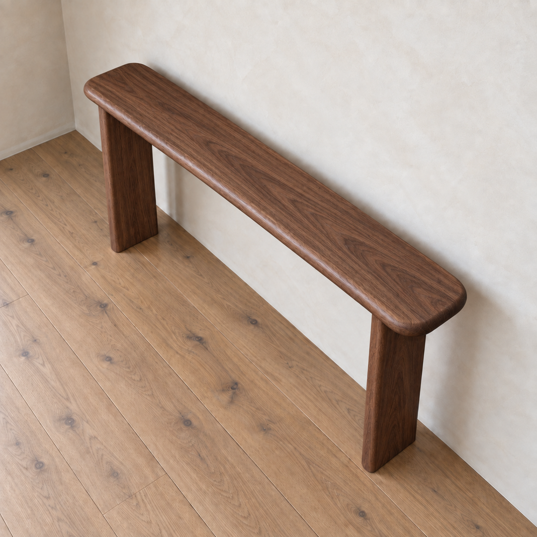 The Narrow console table in walnut features a minimalist, handmade design with rounded edges and two sturdy legs. Crafted from solid walnut for an elegant, timeless touch, it stands against a light beige wall on a wooden floor—perfect as a slim console table in any modern space.
