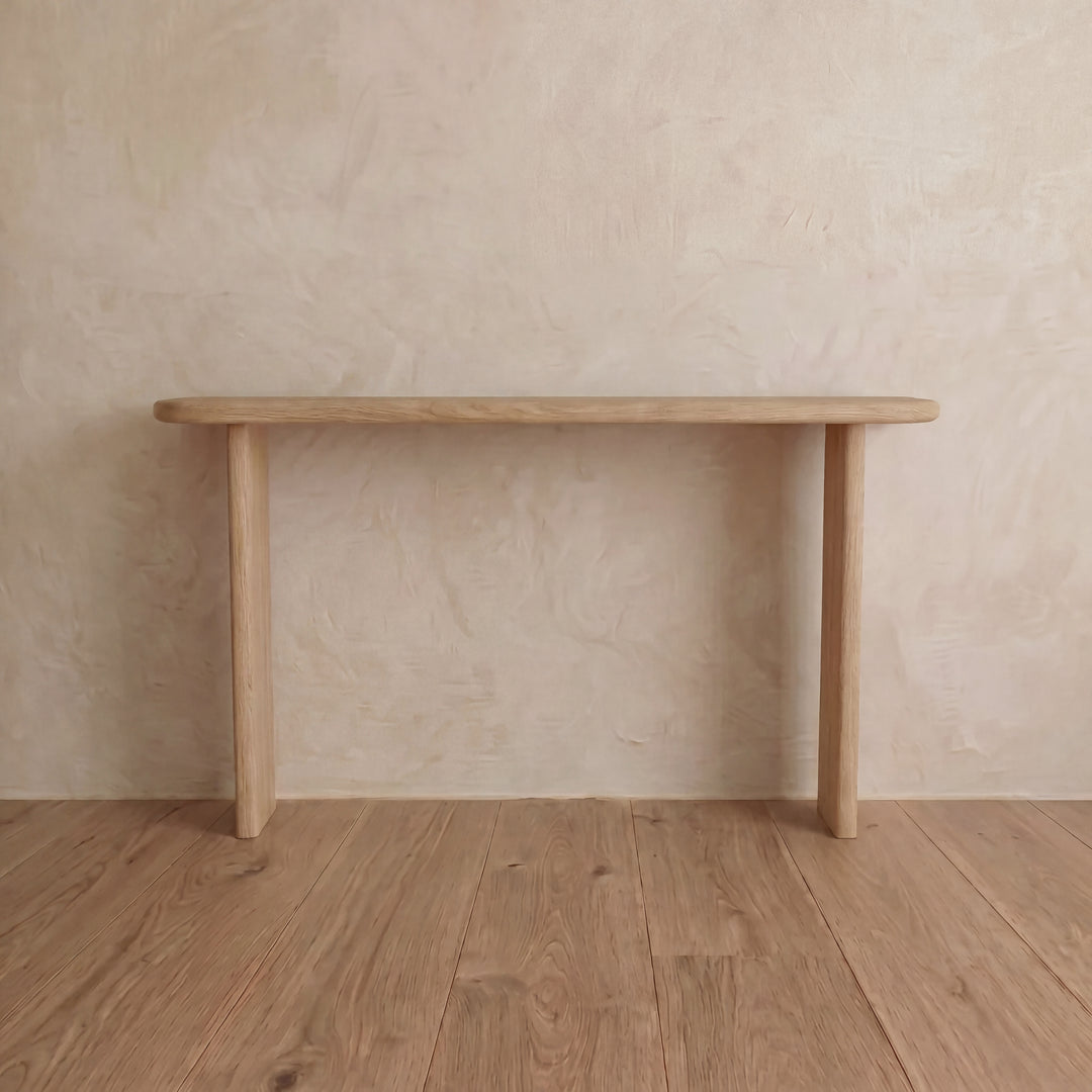 The Narrow console table in white oak features a minimalist design with rounded edges, two legs, and a natural wood finish. Handmade in Vancouver, it stands elegantly against a textured beige wall on a wooden floor.