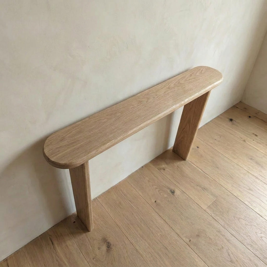 The Narrow console table in white oak, handmade in Vancouver with two end legs, rests against a light-colored wall on a matching floor. Its minimalist, rounded rectangular shape adds understated elegance to any space.