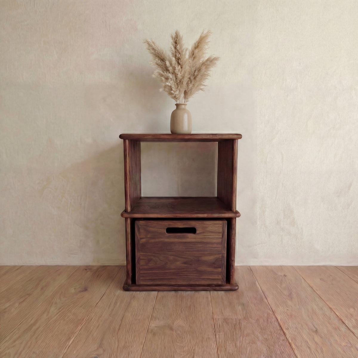 The Nightstand bookshelf in walnut with an open shelf and a storage cube in the bottom opening, topped with a beige vase of dried pampas grass, on a wooden floor against a light textured wall.