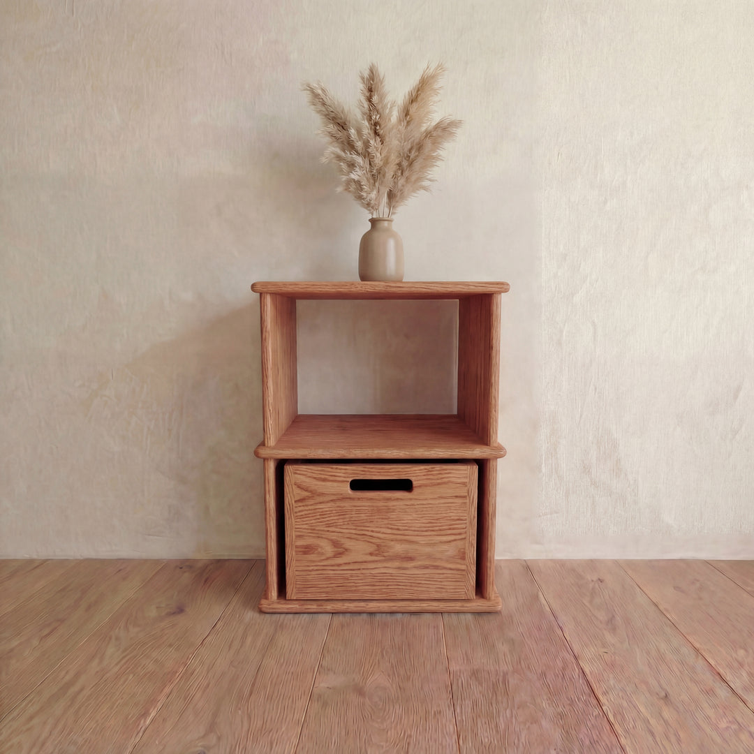 The Nightstand bookshelf in red oak, featuring an open shelf and bottom drawer, is topped with a beige vase of dried pampas grass and sits on a wooden floor against a light textured wall.