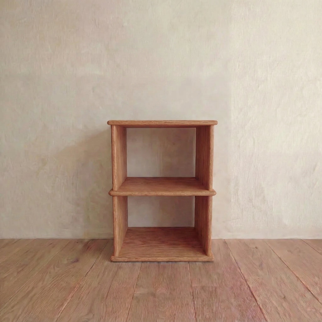 The Nightstand bookshelf in red oak is a small, two-tiered, handmade shelf with an open, box-like and minimalist design, crafted from solid red oak. It’s shown against a plain light wall on a wooden floor.