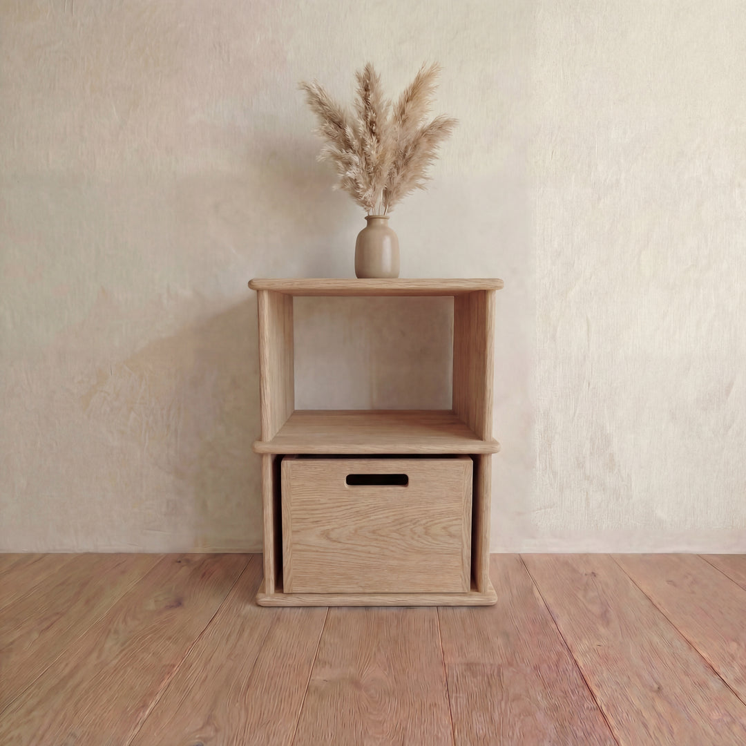 The Nightstand bookshelf in white oak, featuring an open shelf and bottom drawer, stands against a light wall on a wooden floor, topped with a beige vase filled with dried pampas grass.