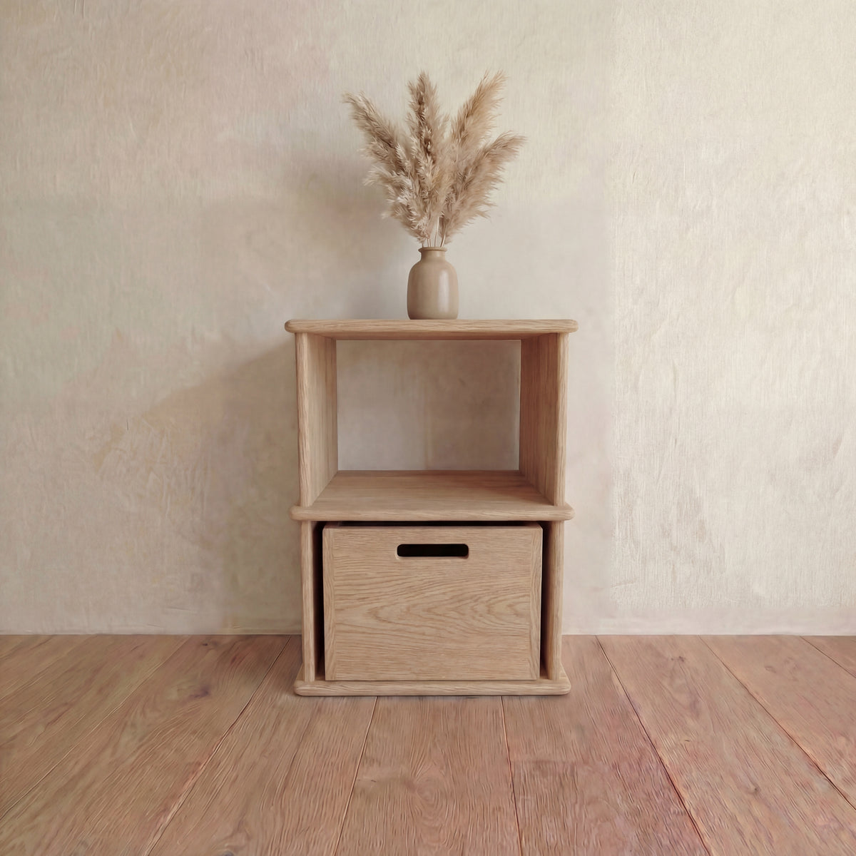 The Nightstand bookshelf in white oak with an open shelf and a storage cube in the bottom opening, against a light wall on a wooden floor, topped with a beige vase of dried pampas grass.