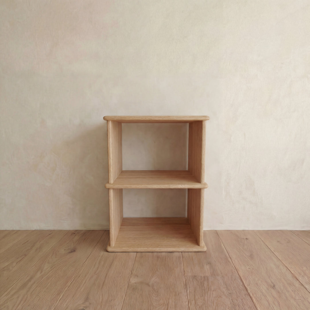 The Nightstand bookshelf in white oak, featuring a simple, minimalist design, stands empty on a wooden floor against a light neutral wall.
