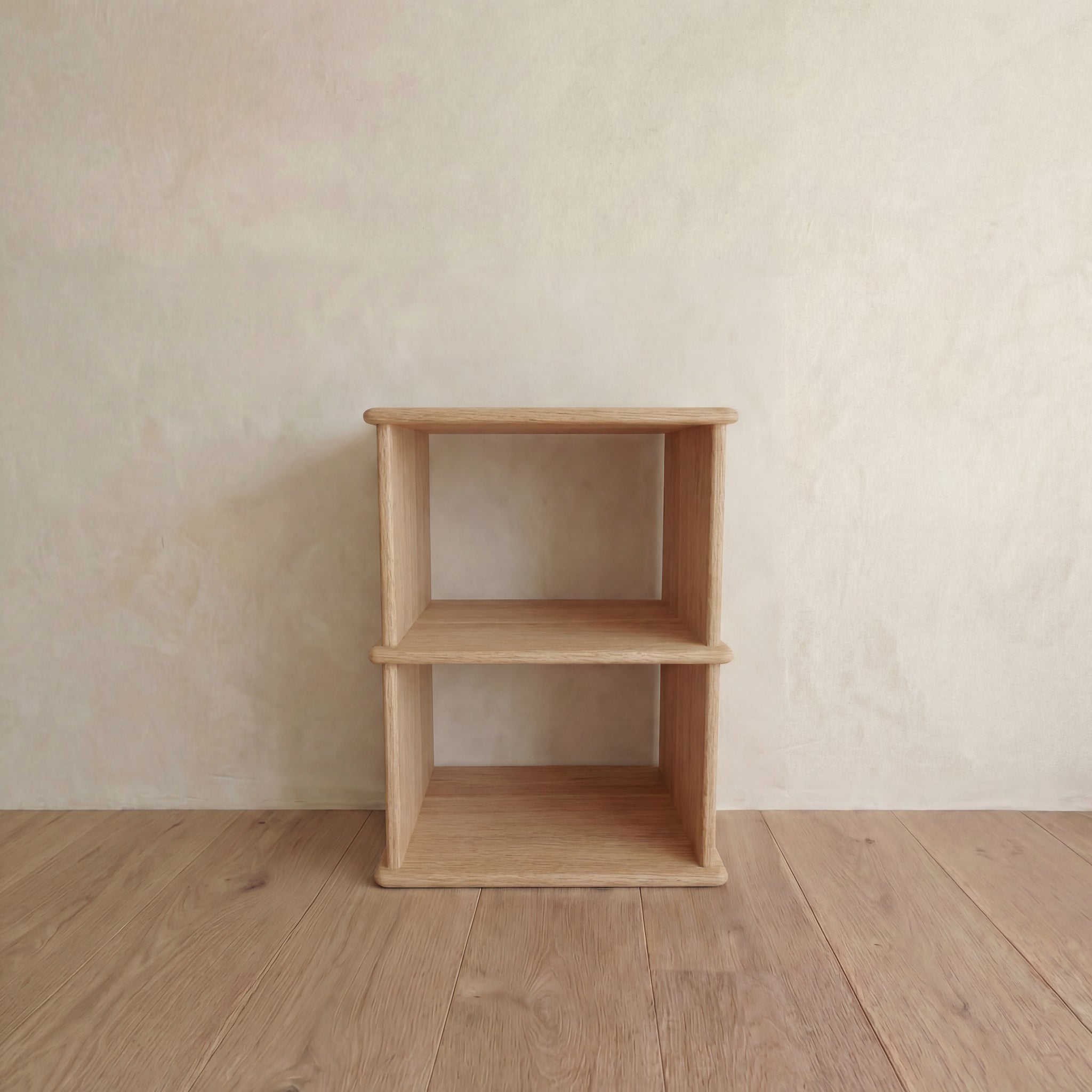 The Nightstand bookshelf in white oak, featuring a simple, minimalist design, stands empty on a wooden floor against a light neutral wall.