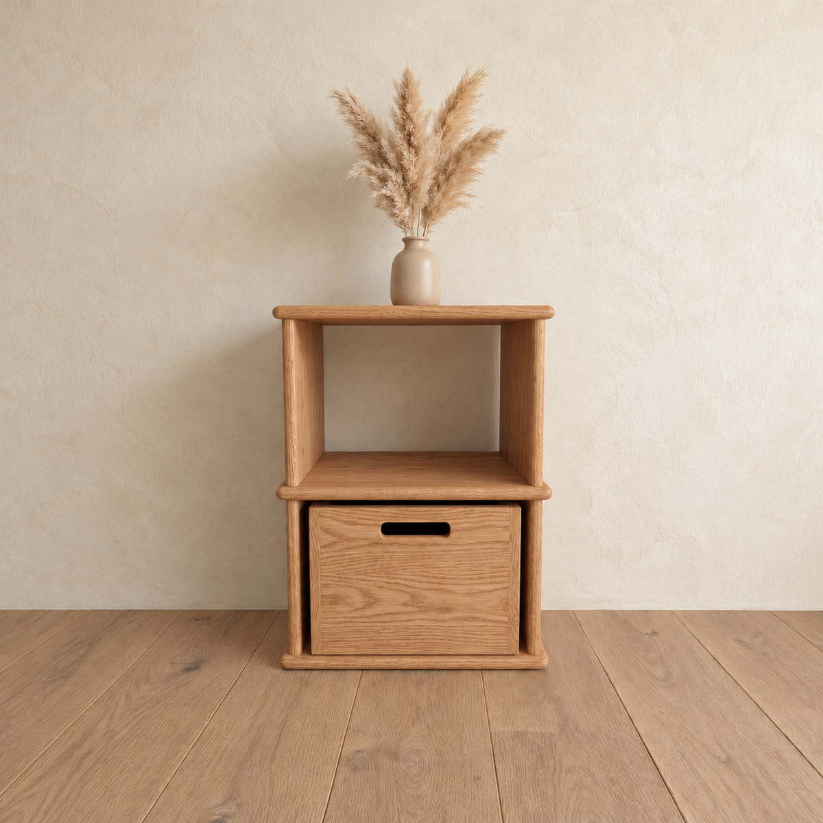 The nightstand bookshelf in red oak against a plaster wall, with a vase of pampas grass on top and a wooden bin in the lower cubby.
