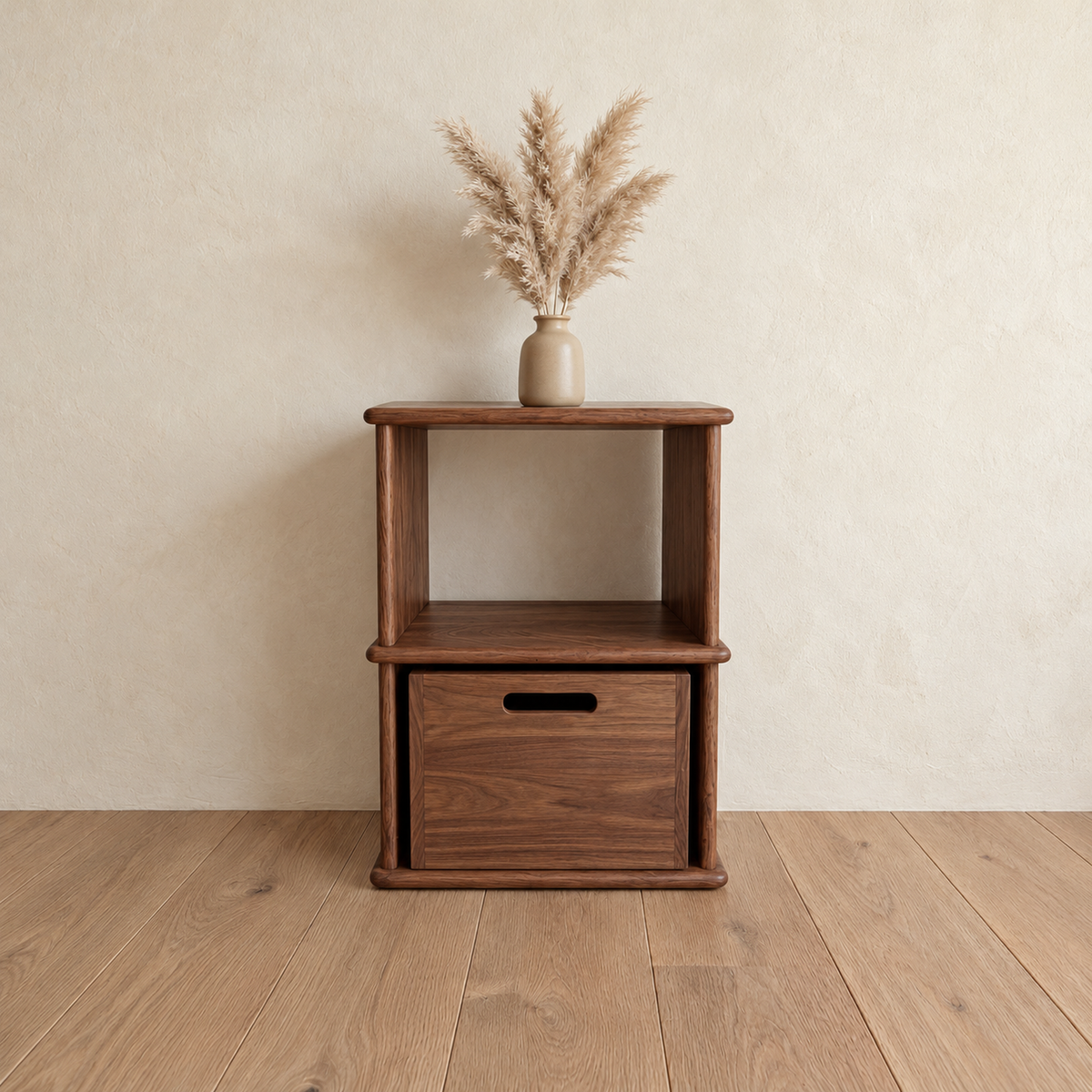 The nightstand bookshelf in walnut against a plaster wall, with a vase of pampas grass on top and a wooden bin in the lower cubby.