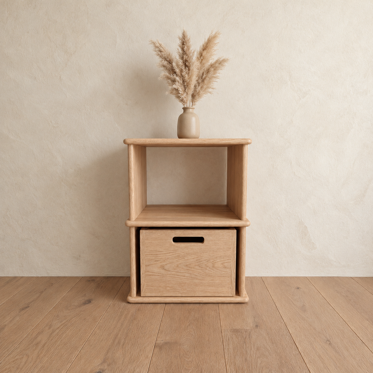 The nightstand bookshelf in white oak against a plaster wall, with a vase of pampas grass on top and a wooden bin in the lower cubby.