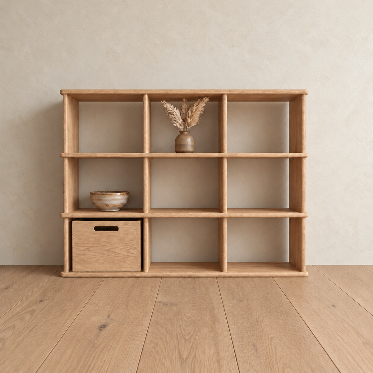 The nine cube bookshelf in white oak against a plaster wall, with a vase of dried grass in the top middle cube, a ceramic bowl on the left and a wooden storage box in the bottom left cube.