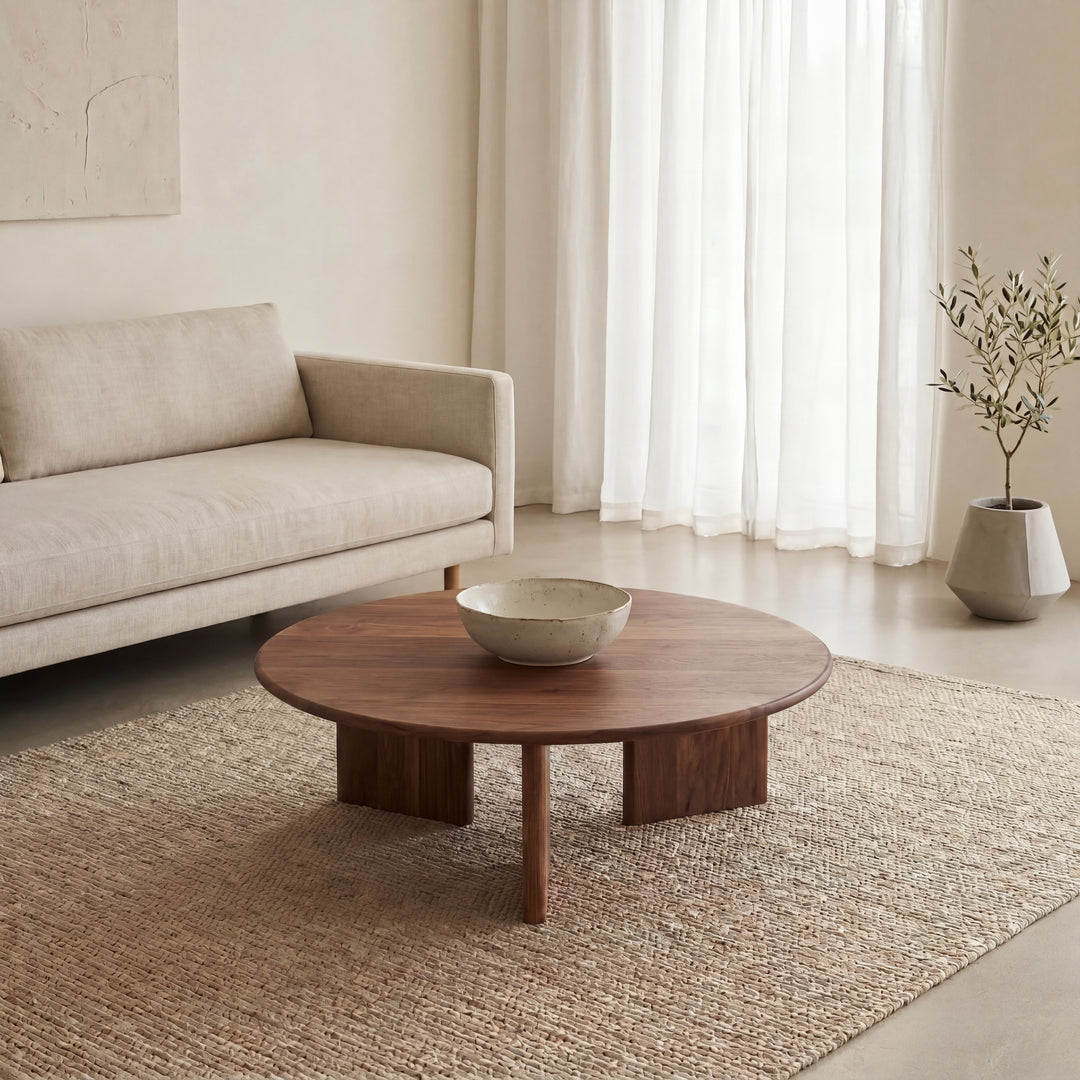 A minimalist living room with a light beige sofa, a Round coffee table in walnut handmade in Vancouver with a ceramic bowl, a woven rug, sheer white curtains, and a potted plant in the corner.