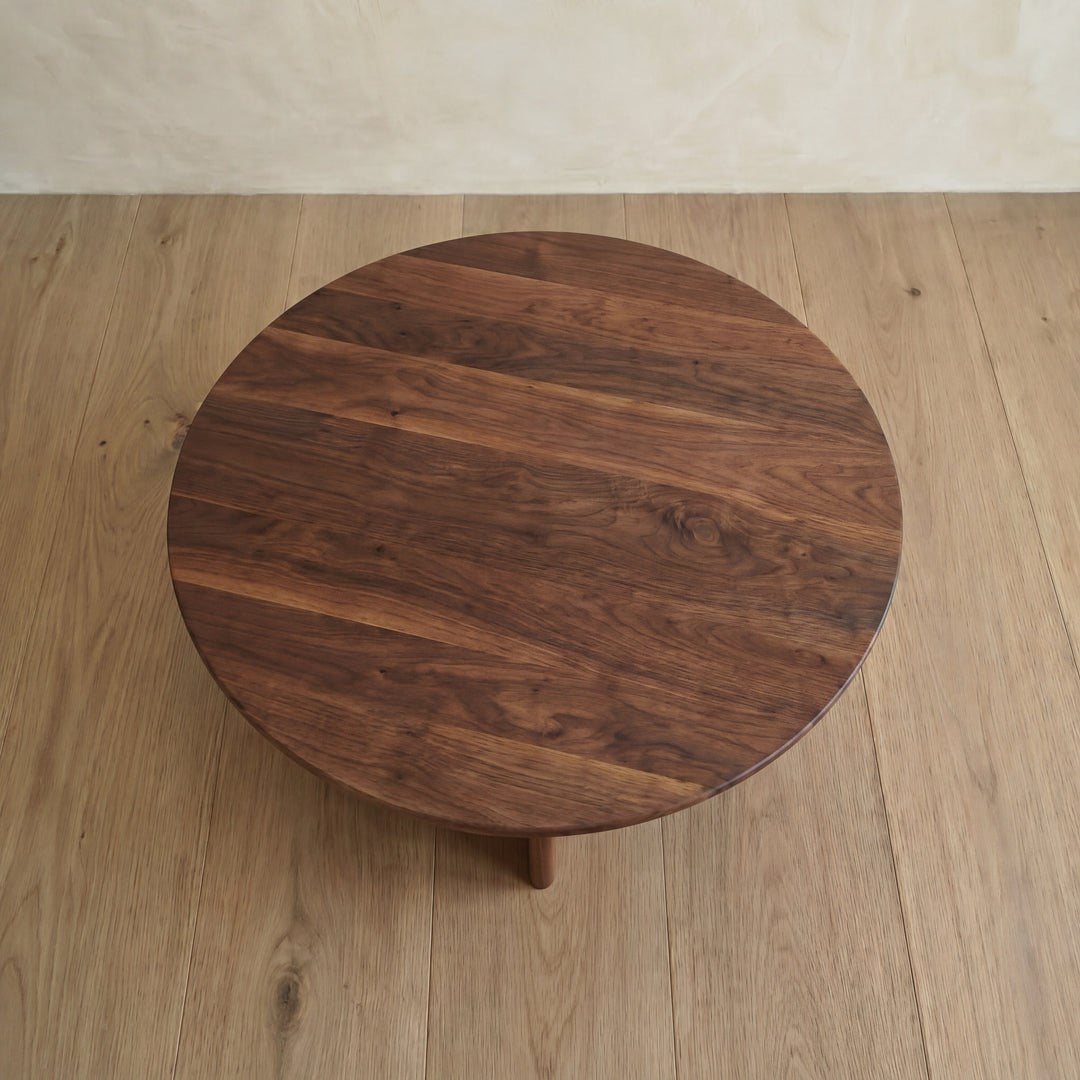 A Round coffee table in walnut, handmade in Vancouver from solid wood with a dark, rich grain, sits on a light wood floor by a beige wall.