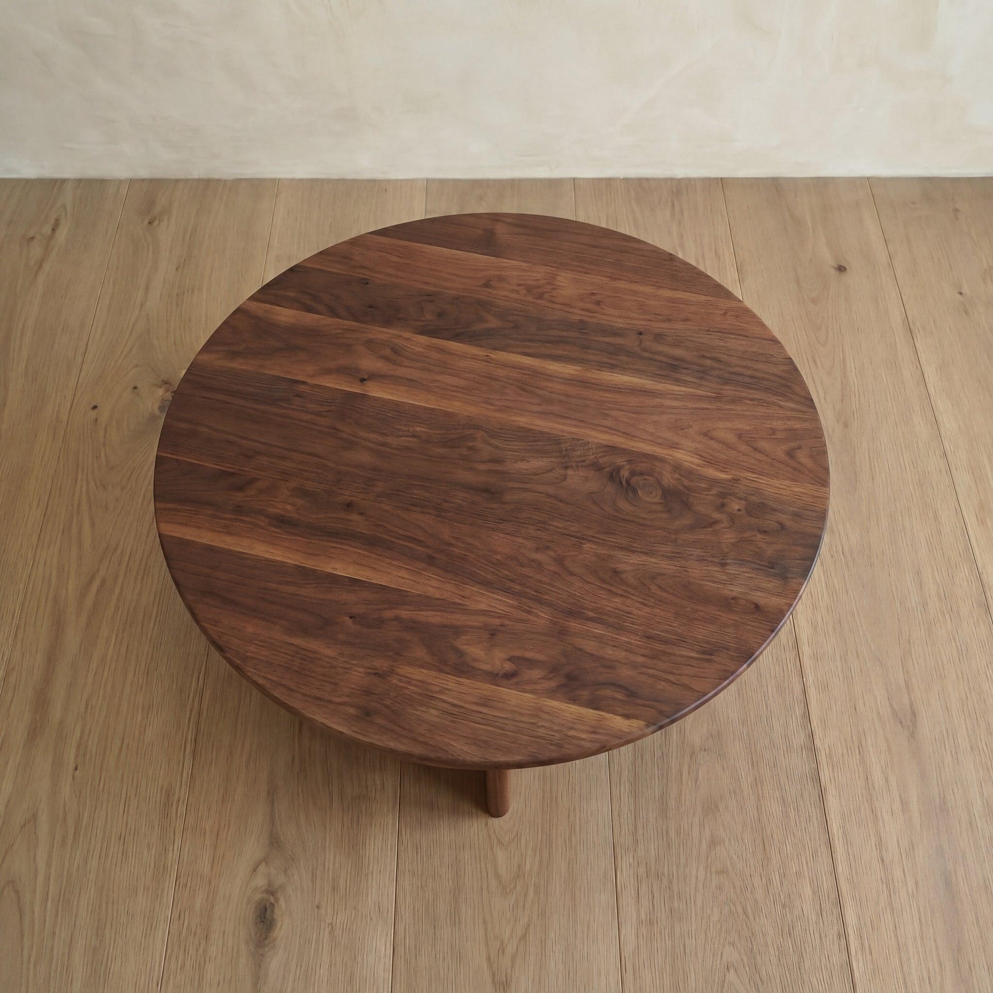A Round coffee table in walnut, handmade in Vancouver from solid wood with a dark, rich grain, sits on a light wood floor by a beige wall.