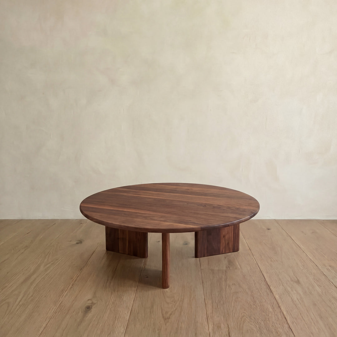 The Round coffee table in walnut, handmade in Vancouver from solid walnut, features a smooth surface and three sturdy legs. It sits on a light wood floor against a beige wall, highlighting its minimalist design.