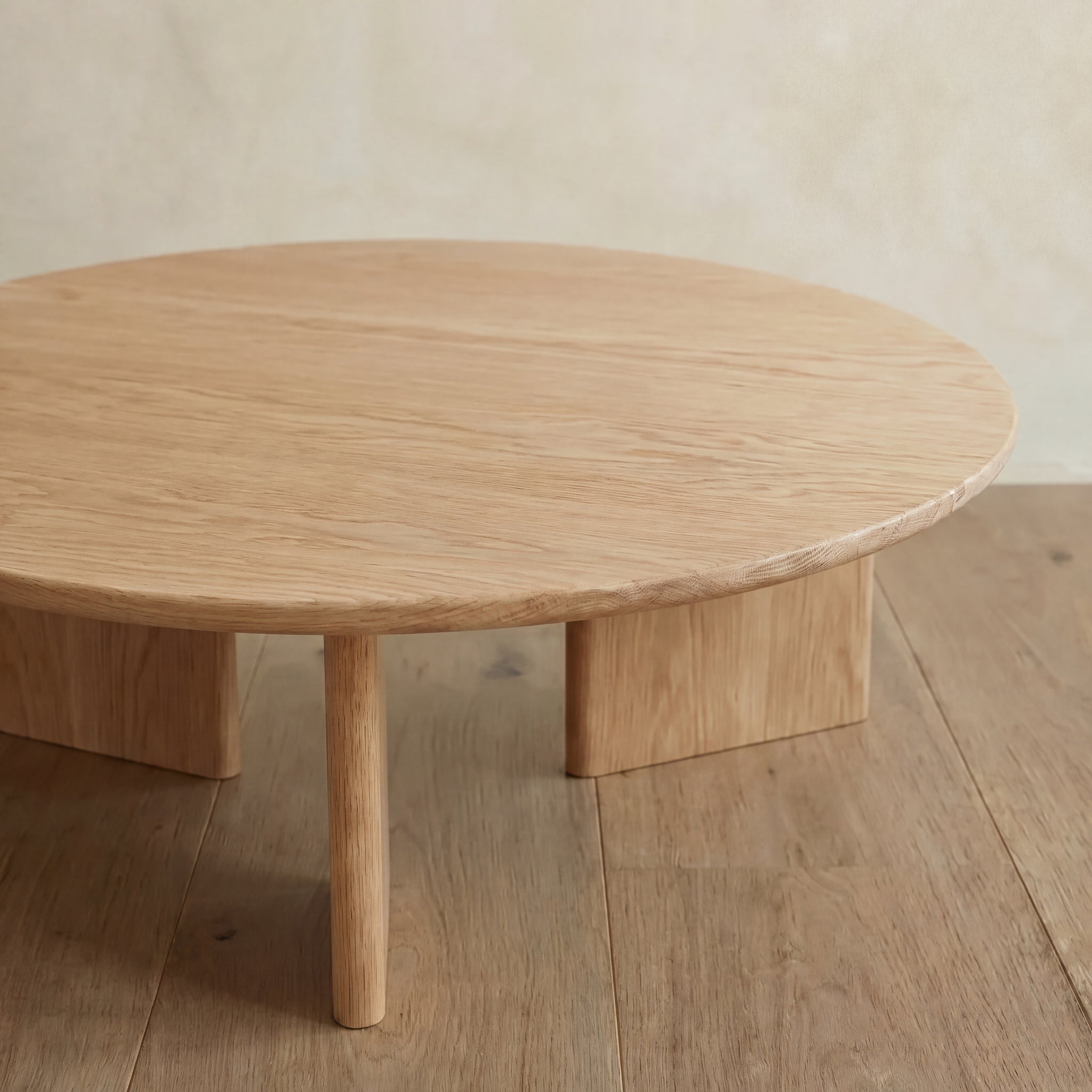 The Round coffee table in white oak features a handmade design with three thick legs, shown on a matching wooden floor against a neutral wall.