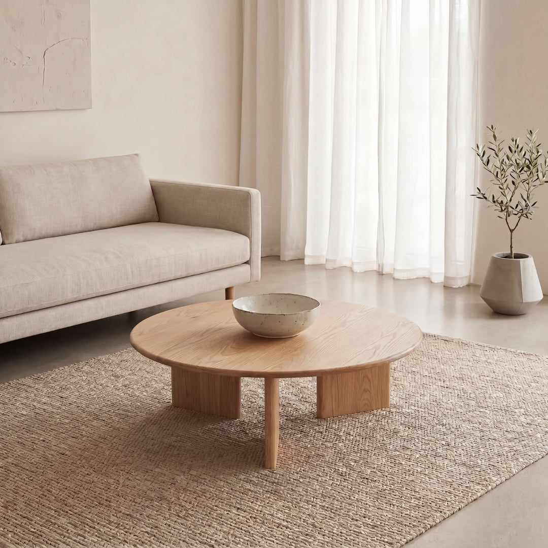 A minimalist living room features a light beige sofa, the Round coffee table in white oak topped with a ceramic bowl, a woven rug, light curtains, and a potted plant in the corner, all in soft neutral tones.