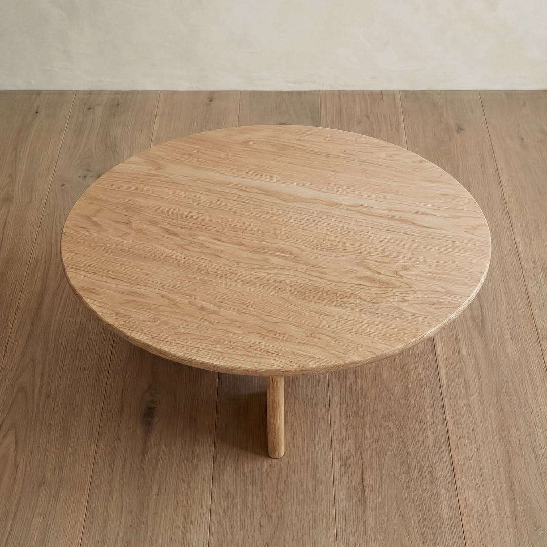 The Round coffee table in white oak with three legs sits on a light wood floor, both featuring similar natural wood grain patterns and light brown tones, as viewed from above.