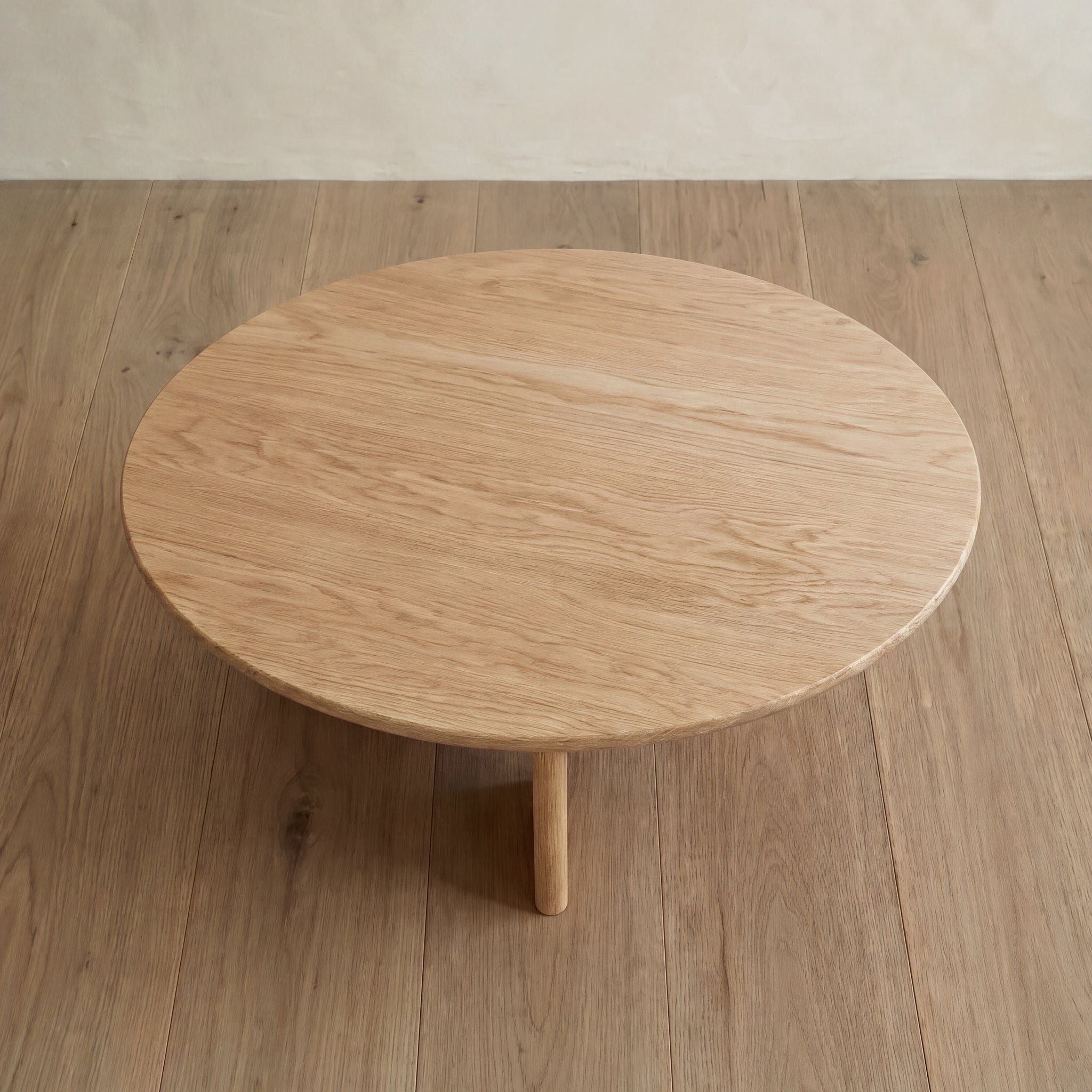 The Round coffee table in white oak with three legs sits on a light wood floor, both featuring similar natural wood grain patterns and light brown tones, as viewed from above.